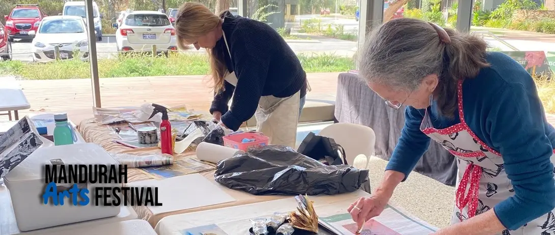 Two women are bent over tables covered with paper, and art supplies, working intently on their art pieces